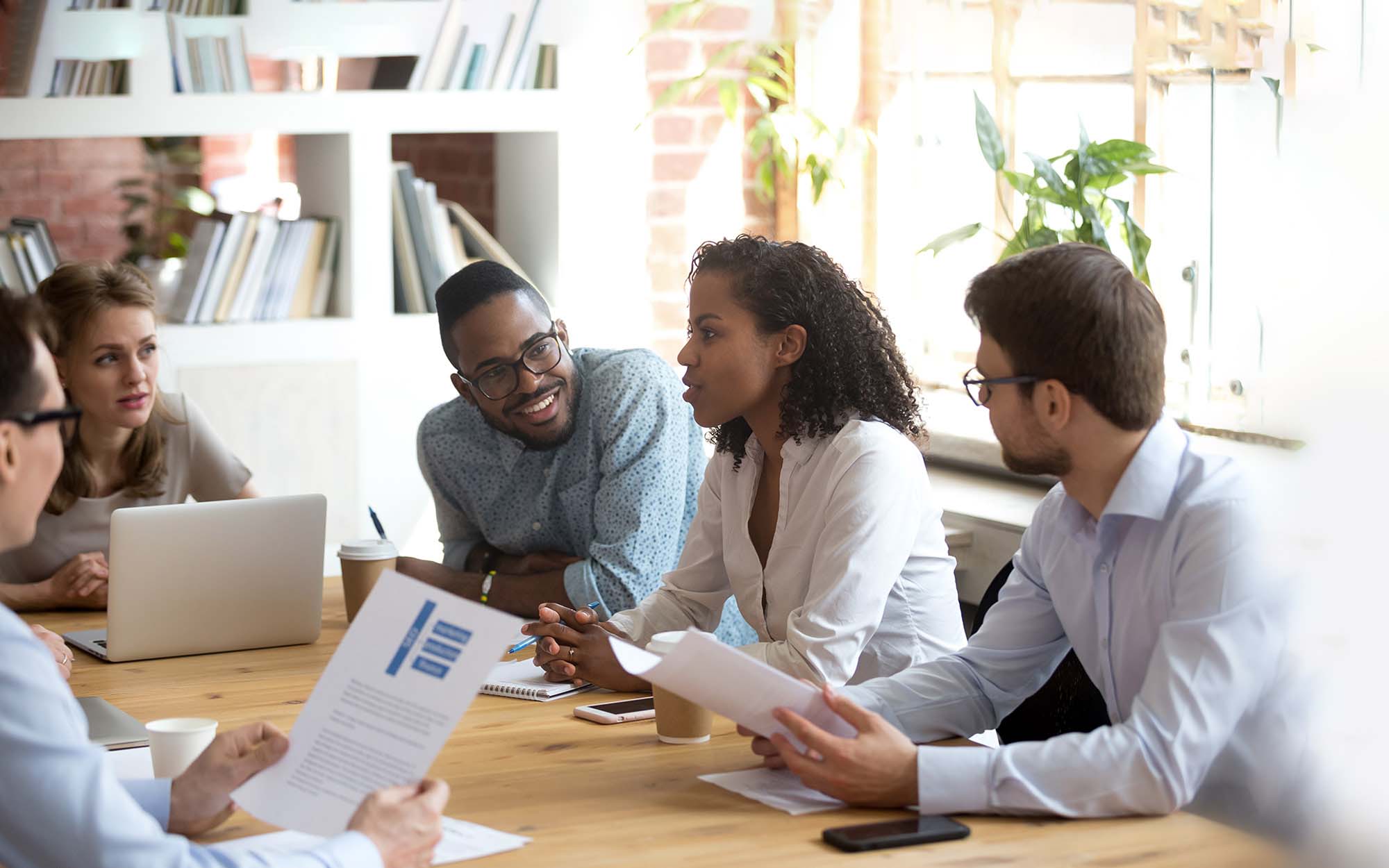 A group of people sit at a table during a meeting holding their notes.